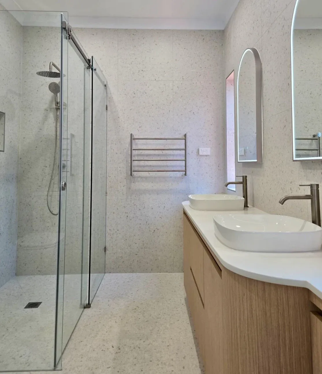 Completed bathroom with curved timber vanity, twin basins, arched mirrors and a glass shower